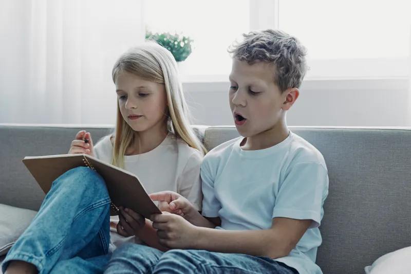 Two children sitting on a couch reading a book together in a bright room. The boy is actively engaged while the girl listens attentively. Concept of education and friendship.