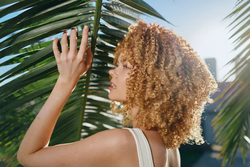 Curly woman touching palm leaves outdoors in sunlight, natural beauty with voluminous hair, warm summer day, close-up profile, vibrant green tropical background.