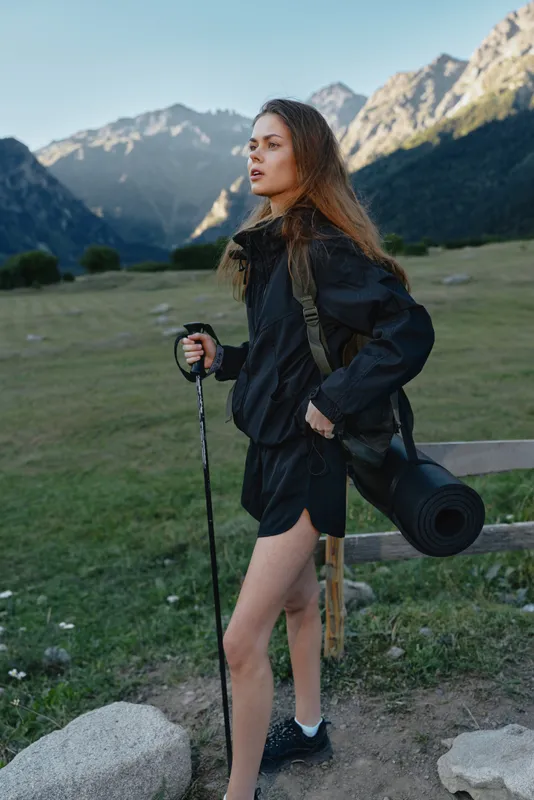 A young hiker stands on a rocky trail in a valley, with towering mountains behind. She wears a dark jacket and shorts, carrying a camera and trekking poles.
