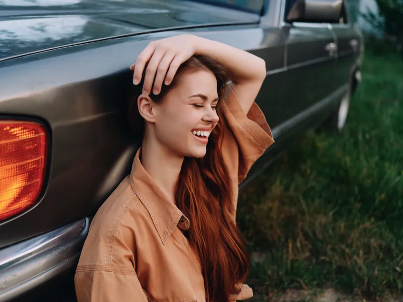 Young woman with long brown hair joyfully laughing near classic car on grassy field, wearing oversized beige shirt, evoking feelings of freedom and nostalgia.