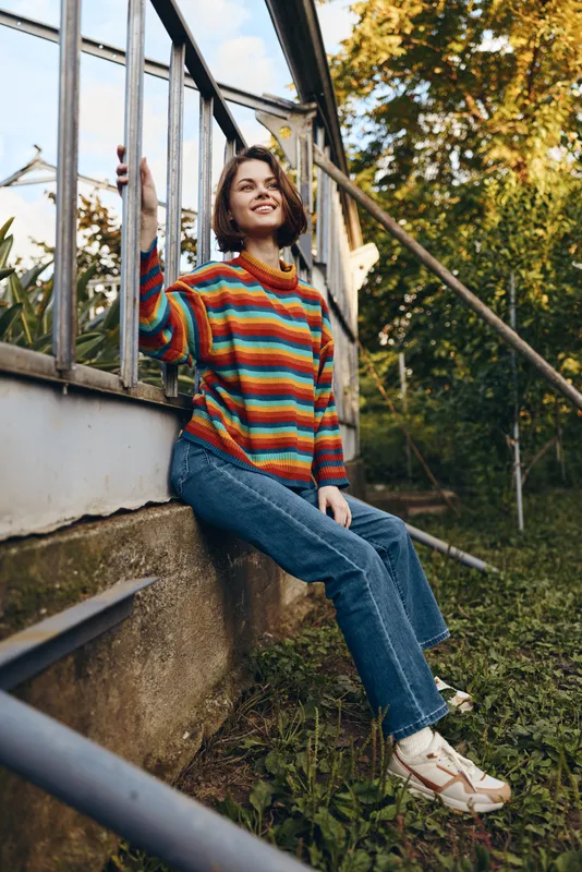 Young woman in her mid 20s wearing a colorful striped sweater and jeans sits leaning on a greenhouse frame in an outdoor garden, smiling warmly in a casual outfit for lifestyle portrait.