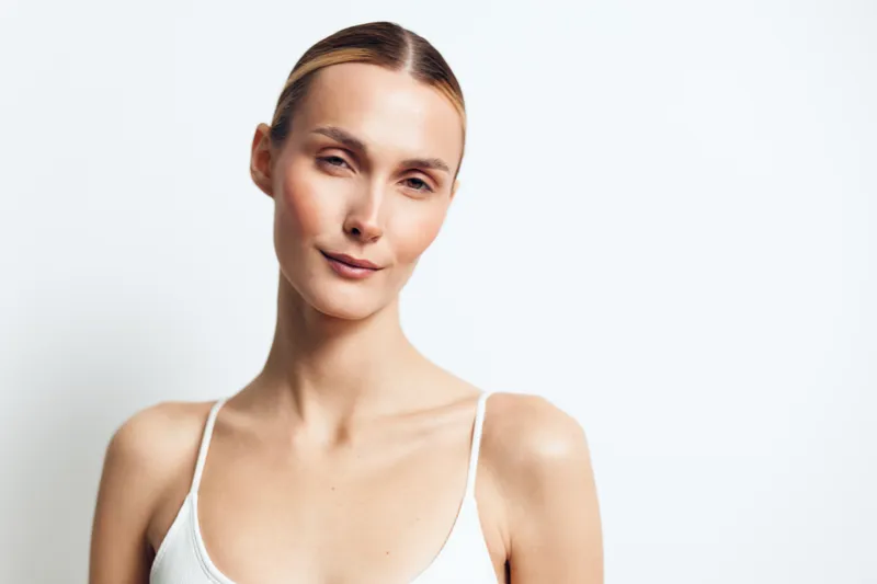 Confident young woman with smooth skin and tied hair wearing white tank top posing against white background with natural makeup and relaxed facial expression in studio light.