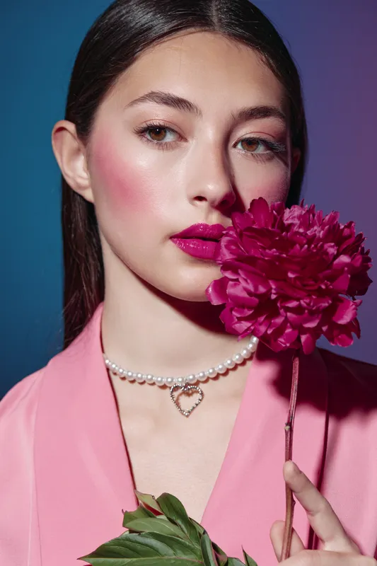 Fashion woman with bold bright makeup and deep pink lipstick holds matching flower near face against blue background created for beauty concept and creative style.