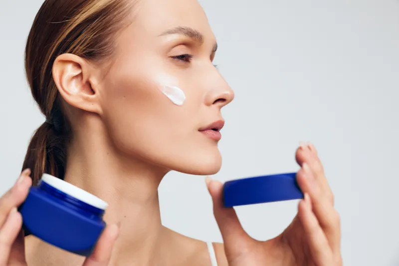 Woman applying skincare cream to face, holding moisturizer jar, focused beauty routine, smooth natural skin, bright studio light, neutral background, self care