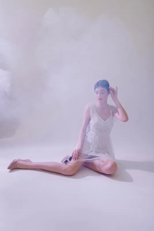 Young woman in white dress sits on floor holding sunglasses in soft light. Concept of beauty, fashion, elegance, and delicate style in minimalistic studio setting.