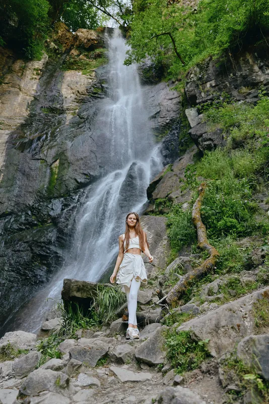 woman standing near a tall waterfall in green forest, wearing casual white clothes, surrounded by rocks and lush vegetation, nature and adventure theme