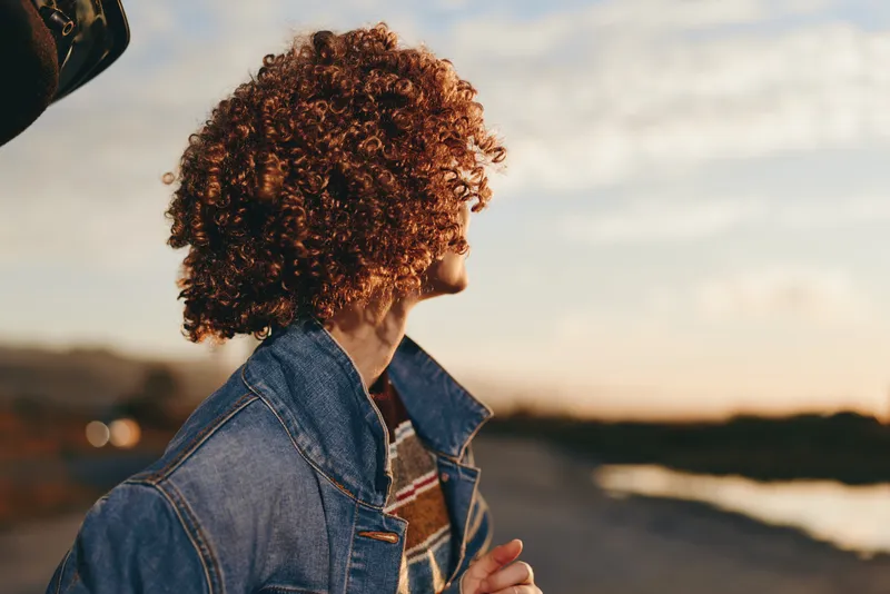 Woman with curly hair, denim jacket, sunset near sea, outdoor lifestyle portrait, side view looking away, warm golden light, relaxed mood, soft bokeh background, warm evening light again