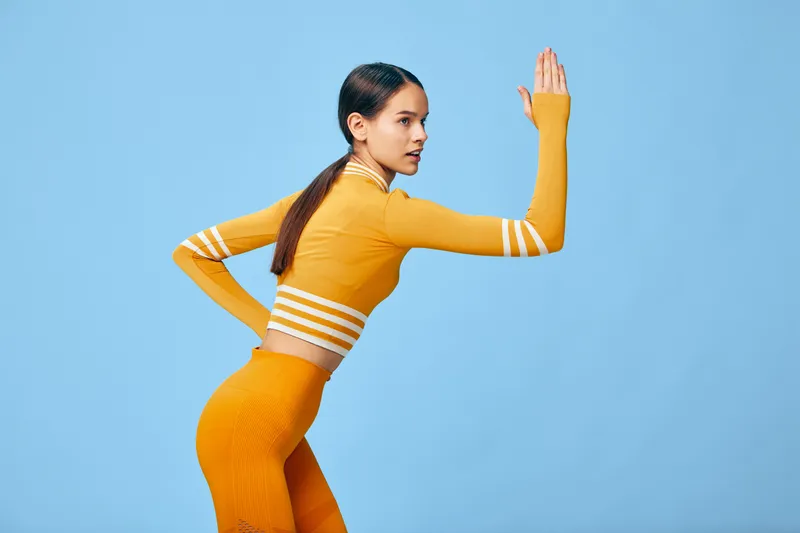 Athletic woman in a vibrant orange fitness set strikes a dynamic exercise pose against a clean blue studio background, highlighting strength, balance, flexibility and modern sportswear aesthetics.