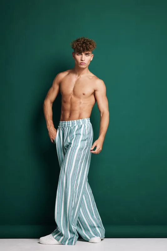 Young man with curly hair posing shirtless in striped wide pants and white sneakers against green background. Fashion, casual style and confident male model standing in studio.