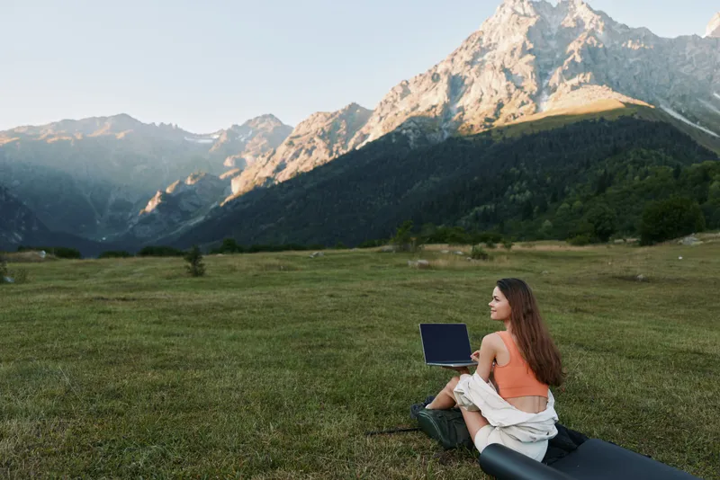 A relaxed woman sits on a grassy field with a laptop, framed by dramatic alpine mountains and a clear sky. She works remotely, smiling as she types, enjoying nature and focus.