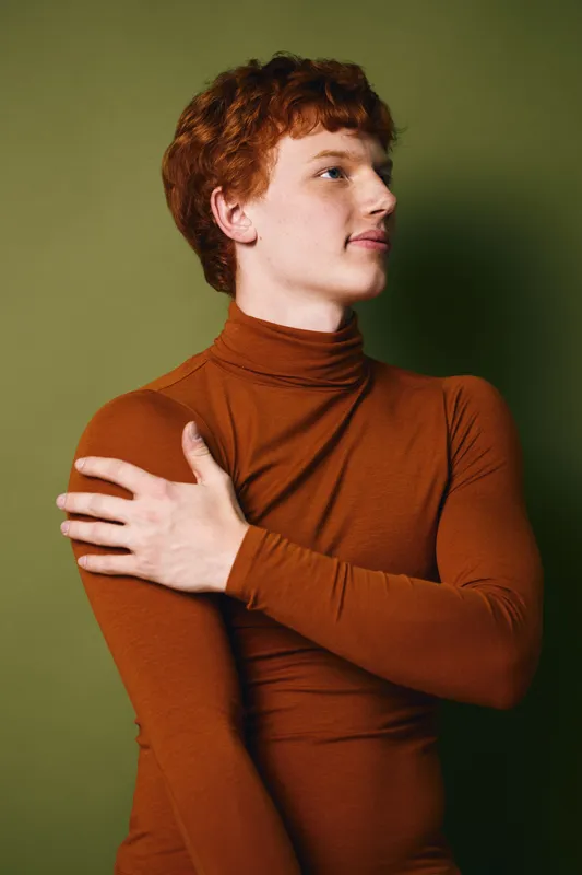 Young man with red hair wearing brown turtleneck sweater standing with hand on his shoulder against green background, posing thoughtfully indoors.