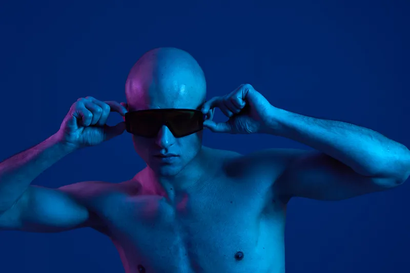 Bold man wearing sunglasses poses under vibrant blue light, showcasing his confidence and uniqueness in a captivating setting. A striking image full of energy.