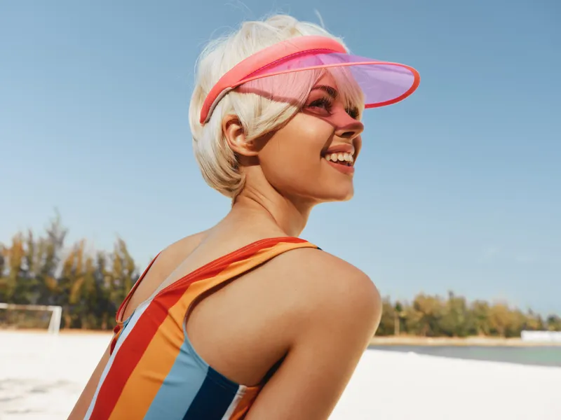Young woman on a sunny beach wearing a colorful striped swimsuit and pink visor, turning with a bright smile against a clear blue sky and calm sea.