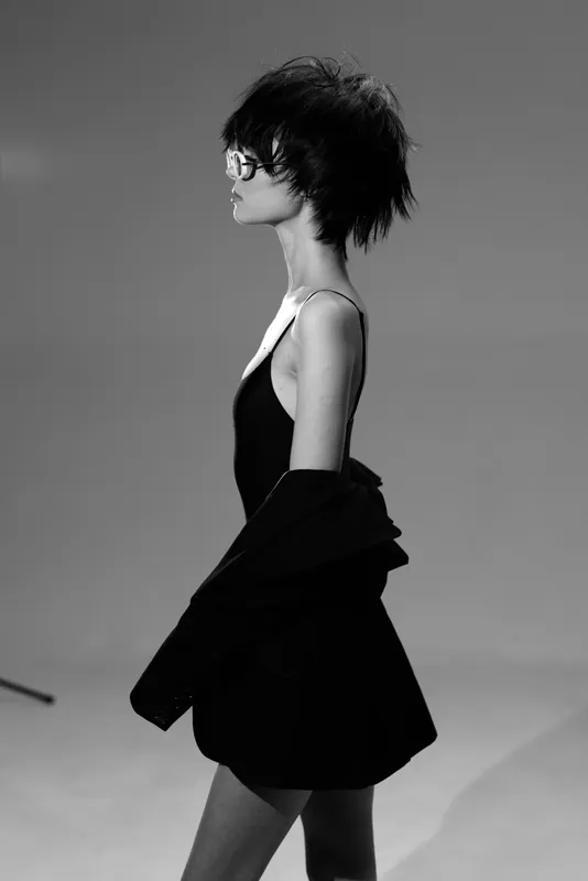 Black and white portrait of a stylish woman with short hair and glasses wearing a jacket and dress in a minimalistic setting.