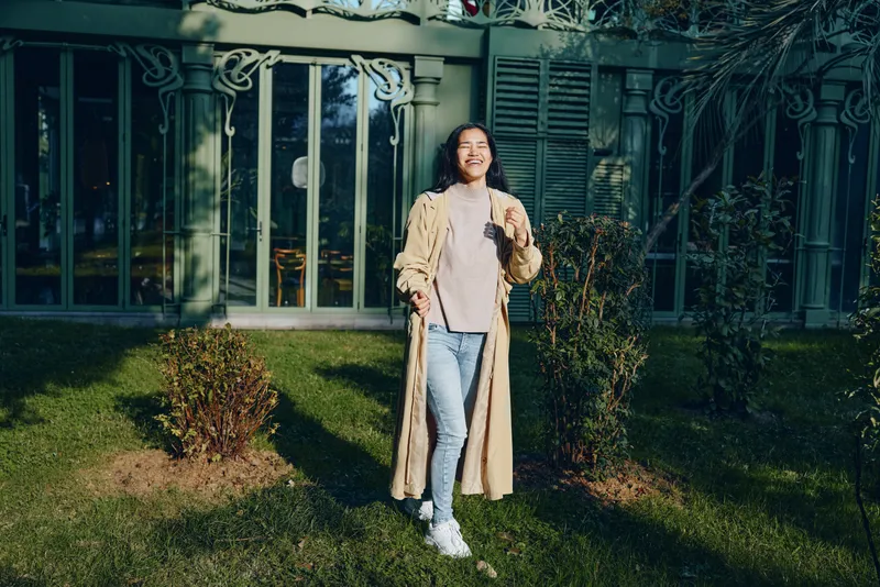 Woman in a beige coat and jeans stands in a sunlit garden in front of a modern house, showcasing casual chic outdoor fashion and a relaxed street style moment