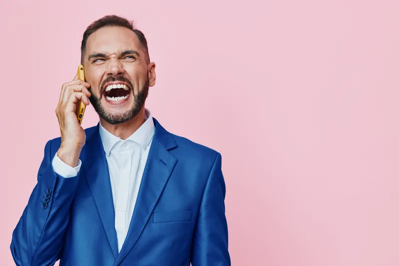 Business professional man on a pink background, shouting into a phone with a tense, energetic expression, isolated colorful backdrop highlighting urgency and bold communication in a modern work scene.