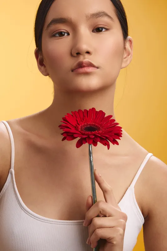 Asian woman holds a vibrant red flower near her face against a warm yellow backdrop, gazing softly at the camera with calm elegance and peaceful mood, inviting quiet reflection and beauty.