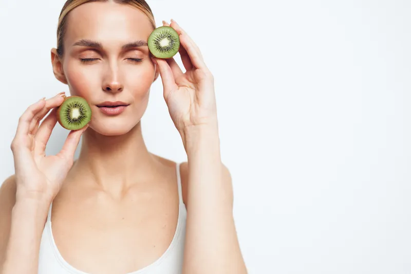 Woman beauty skincare concept, holding kiwi fruit halves near her eyes as a natural face mask, relaxed spa relaxation mood, fresh antioxidant care, soft skin glow, calm portrait on clean background