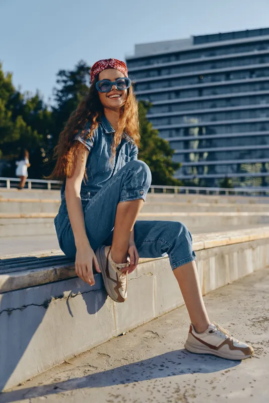 smiling woman in denim jumpsuit and red bandana sitting outdoors on sunny day with sunglasses and sneakers in urban environment