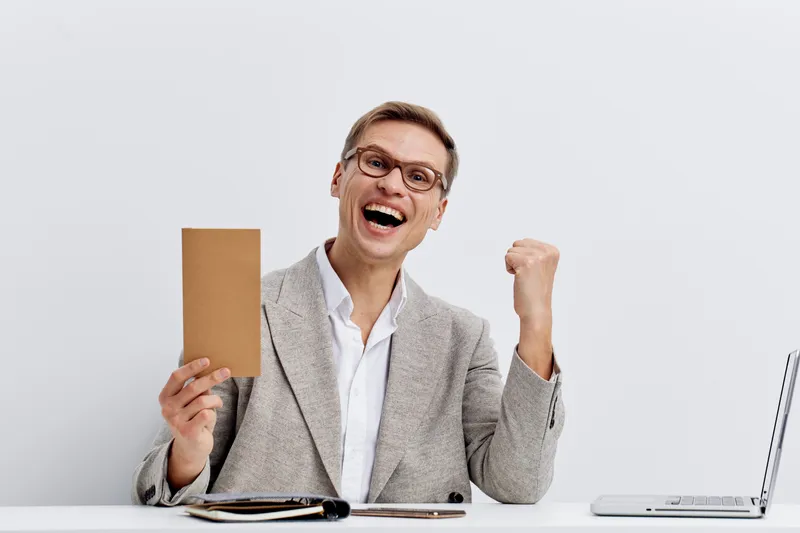Happy cheerful middle-aged man with glasses wearing plaid blazer and white shirt celebrating success holding a cardboard box at his desk studio background. Business achievement concept.