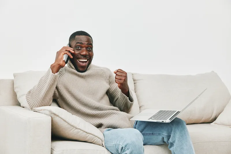 Happy African man celebrating success on a phone call while sitting on a couch with a laptop, wearing a cozy sweater, in a bright and modern living room
