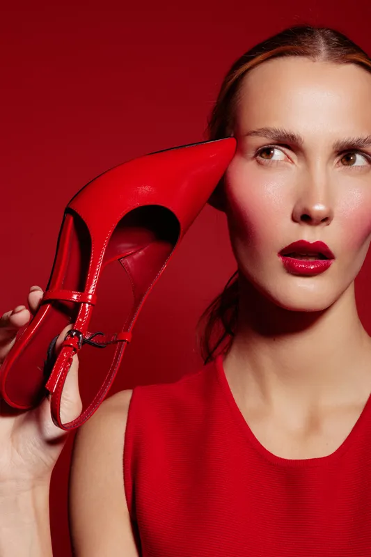 Woman in red dress holds red high heel shoe near face, looking sideways. Bright red background and glossy red lipstick create bold fashion and beauty concept.