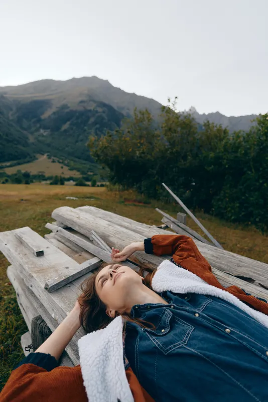 Young woman portrait outdoors in mountains, relaxing on wooden planks in a meadow. Casual denim jacket, eyes closed, peaceful relaxation and rural lifestyle with scenic horizon.