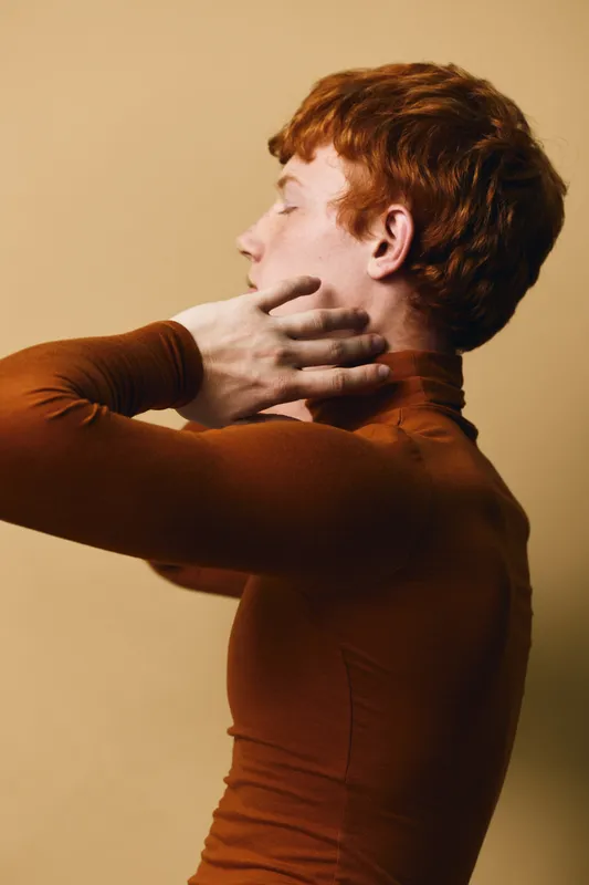 Portrait of young redhead man in brown turtleneck posing with hand on neck looking away on beige background with soft lighting and calm expression in studio.