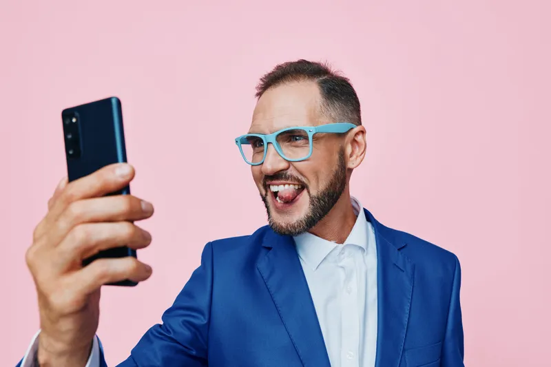 A business professional man poses against a vivid isolated colored background, expressing excitement and confidence as he captures a selfie with a bright smile.