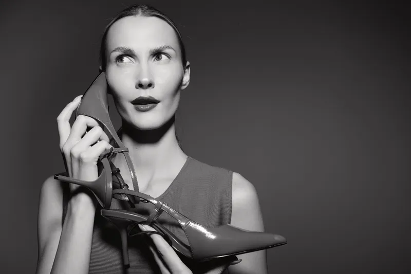 White woman with pale skin holds high heel shoe near face in thoughtful pose against plain dark background. Concept of fashion, style, and elegance.