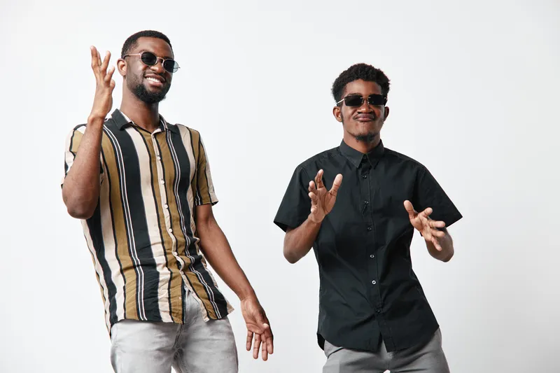 Two young African American men with dark skin wearing sunglasses and casual shirts posing and smiling on white background in studio for lifestyle and friendship.