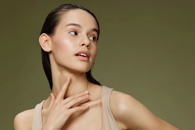 Natural beauty portrait of a young woman with fresh skin, wearing a beige tank top, expressive gaze, and soft lighting against an olive green background, showcasing confidence and simplicity