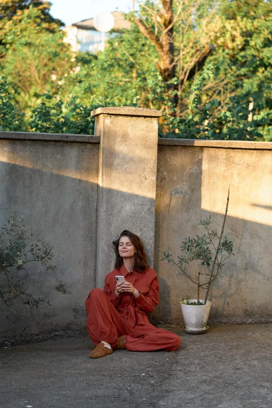 A woman in a red outfit sits on the ground beside a weathered wall, smiling at a smartphone. A relaxed outdoor moment highlighting lifestyle, fashion, and digital connection