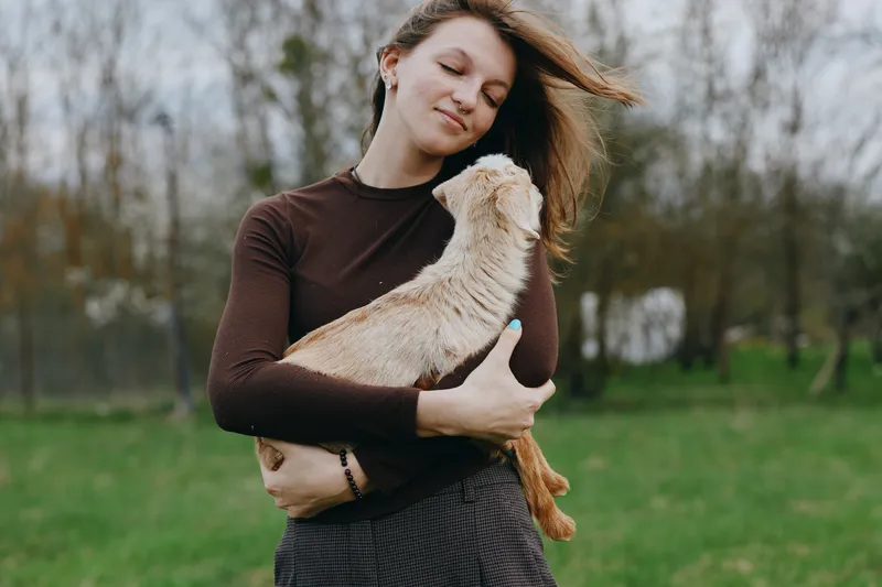 Woman outdoors in a park holds a small dog, smiling softly, wearing a casual sweater, wind in hair, green grass and trees in the background, a warm moment of affection and companionship