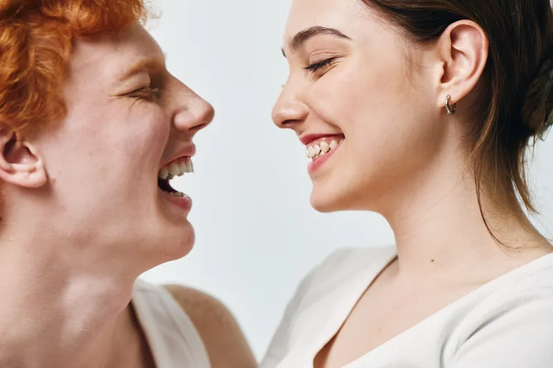 Portrait of two young women smiling and looking at each other in closeup with joyful expressions on light background. Female friends sharing happiness and positive emotions together.