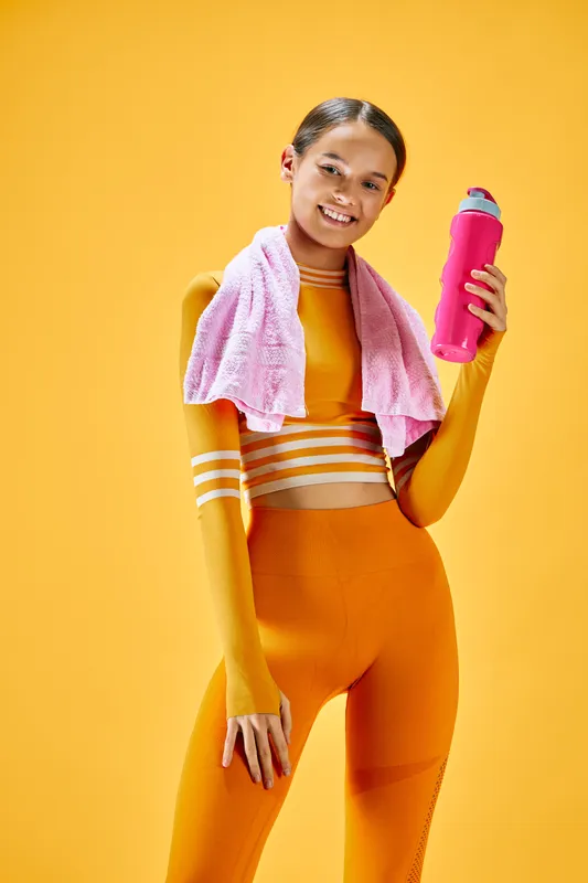 Female fitness enthusiast in a bright athletic ensemble poses with a pink water bottle and towel against a vivid orange backdrop, conveying energy, confidence and active wellness.