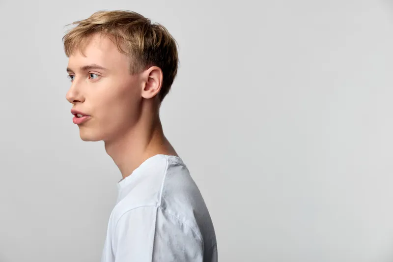 Profile of a young man in a white shirt speaking to the left, studio lighting and a clean neutral background highlight the facial expression and communication.