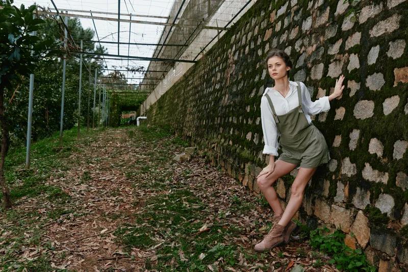 A young woman in an olive green outfit poses confidently against a rustic stone wall, surrounded by lush greenery, evoking a fresh and natural vibe