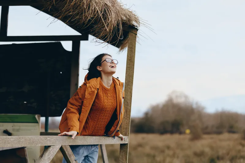 Woman lifestyle portrait outdoors in winter, smiling while leaning on a wooden porch rail, wearing an orange jacket and scarf, looking upward in sunlight, countryside field landscape behind, calm mood