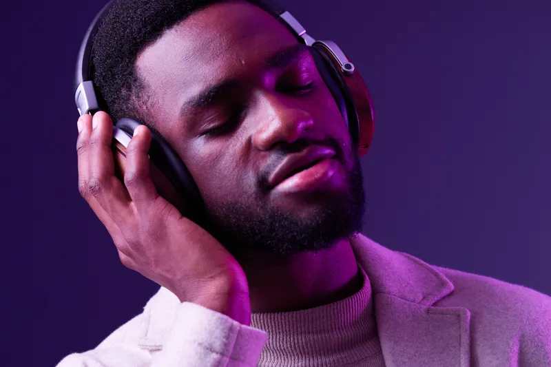 Young Black man enjoying music with headphones against a purple background, showcasing relaxation and emotional connection through music, modern fashion style