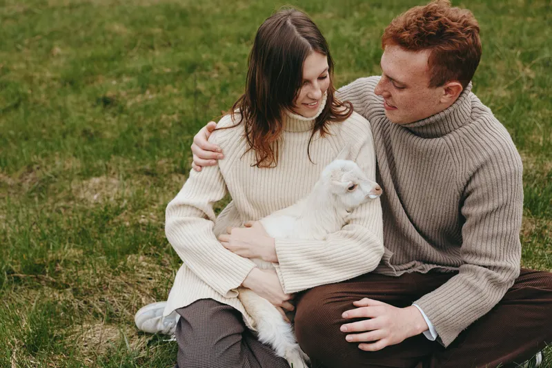 A couple sits on the grass outdoors with a small goat kid, sharing a gentle moment of affection and warmth. A calm, rustic scene highlights companionship and family life in nature.