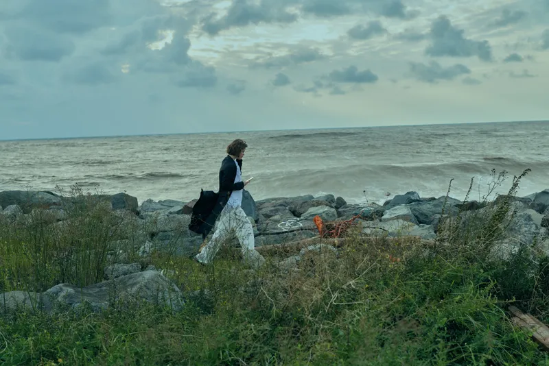Woman walking along a rocky coast with a bag, sea on the horizon, and a cloudy sky, conveying outdoor travel, nature, and coastal exploration today.