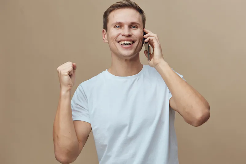 smiling young caucasian man talking phone celebrating success white t-shirt beige background digital nomad concept