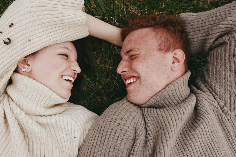 Smiling couple lying on grass wearing chunky knit sweaters, enjoying a cozy outdoor moment. warm natural tones create a relaxed lifestyle scene focused on affection, comfort, and shared laughter.