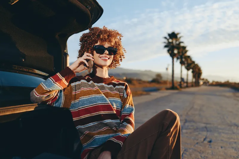 Woman lifestyle, curly hair, sunglasses, striped sweater, sitting by car, talking on phone on roadside, palm trees and landscape at sunset, warm light, relaxed casual travel mood
