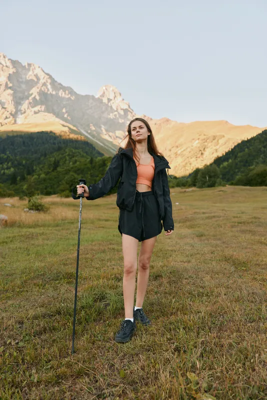 A confident outdoor enthusiast stands in a grassy alpine meadow with a trekking pole, wearing a jacket, shorts, and boots. Sunlit mountains rise behind, creating a vivid adventure scene.
