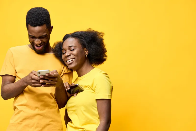 Happy young African couple using smartphones together against a bright yellow background, showing joy and connection in a cheerful, vibrant setting