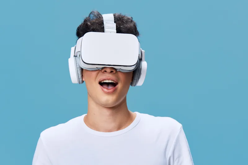 Smiling man wearing virtual reality headset and wireless headphones on blue background, enjoying immersive technology with casual white t-shirt, happy digital experience.