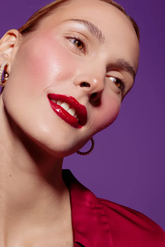 Close-up portrait of a smiling woman with glossy red lips and radiant skin wearing hoop earrings and a red blouse. Studio lighting highlights her natural beauty on purple background.