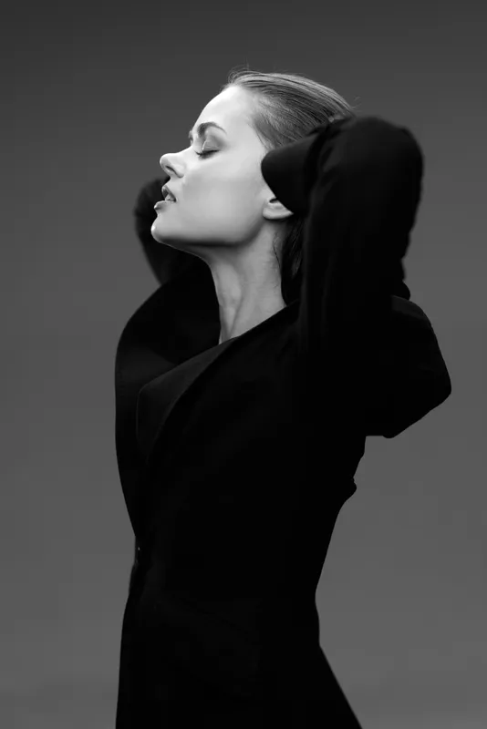 Black and white portrait of a thoughtful woman wearing a black coat with closed eyes and arms raised.
